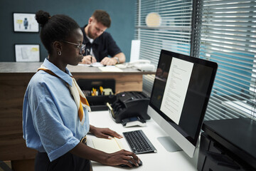 Young Black woman working at computer in bank office, standing at desk and typing on keyboard while middle aged Caucasian man writing in background, modern workplace setting