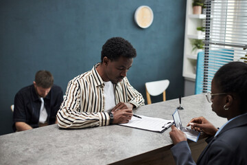 Black young man filling out paperwork at bank office counter while Black female clerk holding identification card assisting him, Caucasian male customer sitting in background waiting