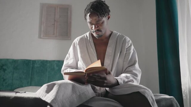 Man in bathrobe sitting on bed, reading book with focus. Bright morning light fills room, soft atmosphere. Quiet, cozy indoor scene with tranquil mood and relaxed vibes