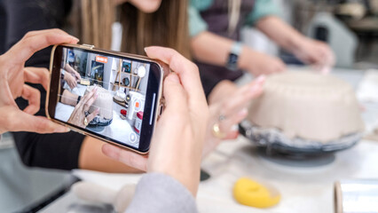 In an art studio with  a beautiful middle-aged woman artist is shaping clay at a table, while a young woman next to her is taking a photo with her phone