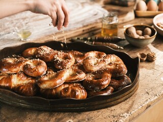 Hand sprinkles crushed walnuts atop golden brown, freshly baked Mucenici arranged on a wooden tray, with eggs and honey in the background, ready to be enjoyed