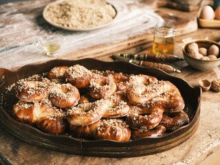 A rustic wooden table holds a platter overflowing with golden Mucenici, sprinkled with walnuts. Baking ingredients hint at the creation of this delightful Romanian treat