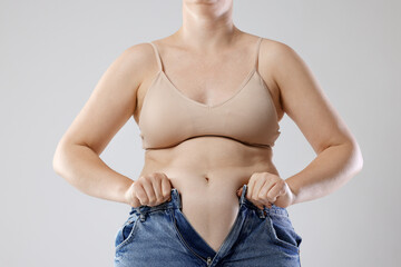 Liposuction. Woman trying to put on tight jeans against grey background, closeup