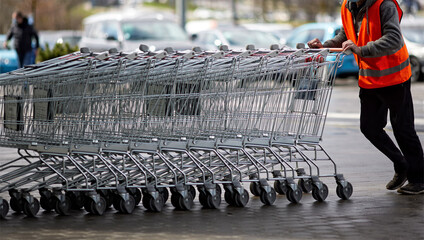 Supermarket employee in orange vest pushes multiple empty metal trolleys across parking lot. Stacked wire carts ready for customer use during retail activities. Labor, service industry, routine work