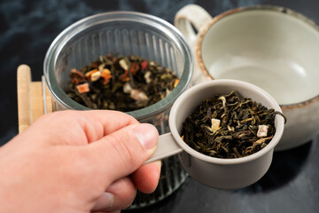 A hand holds a measuring cup filled with loose leaf tea, ready to pour into a tea infuser. A glass container with more tea sits nearby, creating a relaxed afternoon atmosphere in the kitchen.