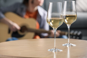 Glasses with white wine on table and man playing guitar indoors, selective focus