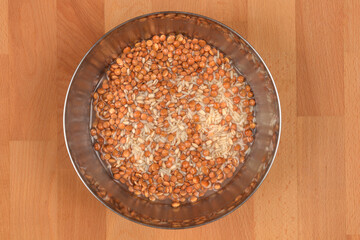 An overhead shot of a metal bowl filled with soaking brown lentils, barley, and rice on a wooden surface
