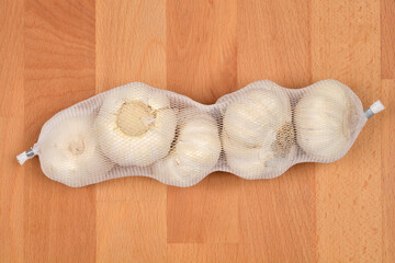 A string of fresh garlic bulbs in a white mesh bag is photographed on a wooden background from a top-down perspective