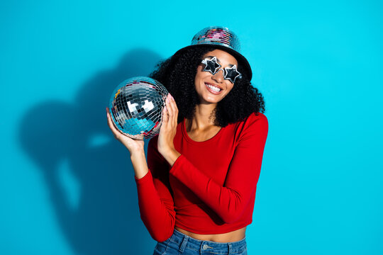 Cheerful young woman holding a disco ball, wearing a red top and star shaped sunglasses, against a bright blue backdrop