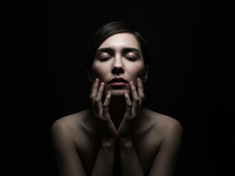 A low-key portrait of a woman with closed eyes and hands framing her face against a dark background, evoking a sense of calm and introspection.