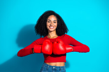 Confident young woman with boxing gloves posing against a vibrant blue background, expressing strength and positivity.