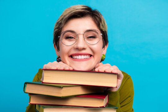 Young stylish woman with short hair wearing khaki shirt leaning on stack of book smiling against blue background