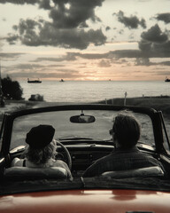 Rear view of two people sitting in a convertible car, facing the sea at sunset with cargo ships on the horizon under a partly cloudy sky