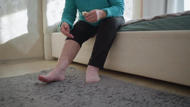 Old woman in green applying ointment to her hand while sitting on the edge of the bed. One sock on, the other off, as she takes care of herself in a cozy, peaceful bedroom setting with soft light