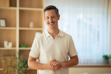 Smiling middle-aged man standing indoors with folded hands, looking confident and friendly in a bright modern home environment.
