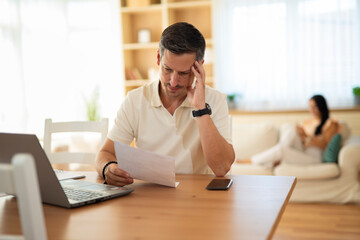 Stressed man reading financial document while working from home, feeling worried about expenses.