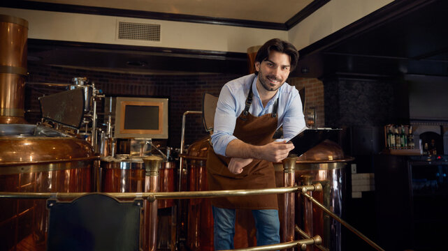 A man wearing an apron is studying a clipboard in a brewery setting - Powered by Adobe