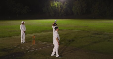 Diverse male cricket teammates pausing, bowling, inspecting ball and questioning delivery at night