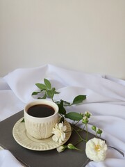 Minimalist morning scene with coffee cup, blooming white roses and closed journal on draped fabric.