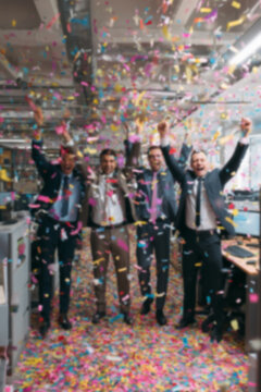 Blurred background of people in business suits celebrating in an office, throwing confetti and holding party streamers. Concept of corporate celebration, team success, and joyful workplace moments.