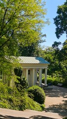 Small classical building with white columns surrounded by lush green trees in a park. Concept of architecture in nature, historical charm and peaceful outdoor space