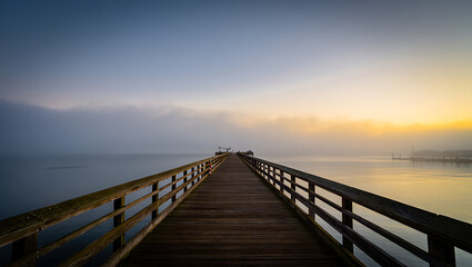 A wooden pier stretches out into the calm ocean under a gradient sky of blue and golden hues at dawn or dusk, with a gentle mist hanging over the water