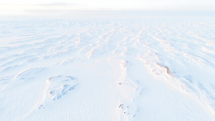 An ethereal winter landscape unfolds with gentle snowdrifts creating a mesmerizing pattern across the vast, frozen terrain under a pale, hazy sky