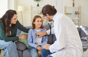 Obraz premium Friendly male doctor in white coat using stethoscope, listening to heartbeat of smiling child during medical examination. Pediatrician making house visit to mother and kid for health examination.