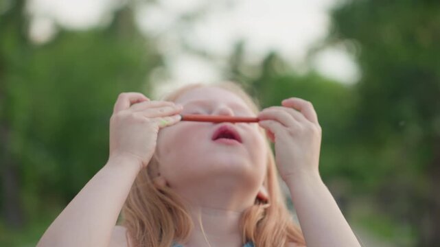 close up young girl holding paint brush under nose sniffing colorful paint playfully with eyes closed and lips puckered while seated outdoors by water against blurred green trees in warm evening