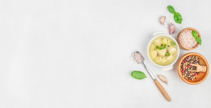 Garlic cloves marinated in olive oil with basil and pepper, served in a white bowl, next to bowls of pink Himalayan salt and colorful peppercorns, on a white background. copy space. - Powered by Adobe