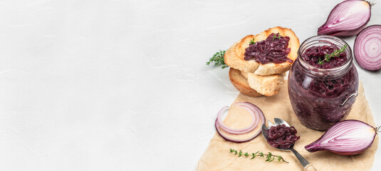 Glass jar of homemade caramelized red onion jam with thyme, served with toasted sesame bread slices, fresh red onion halves, and rings, on a white background, copy space