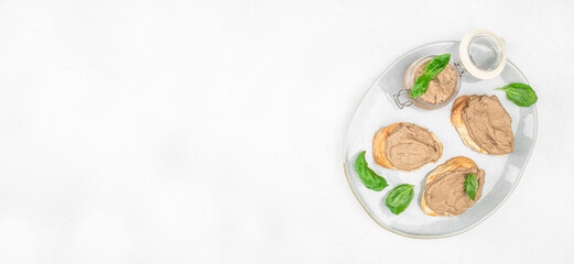 Jar of creamy pate with fresh basil, served on toasted bread slices, arranged on a white oval plate over a light background copy space