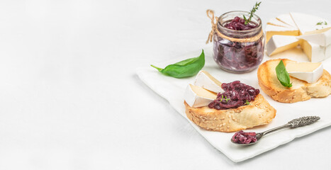 Brie cheese with caramelized red onion jam on toasted sesame bread slices, served with fresh basil, a jar of onion chutney, and a spoon, on a white marble board with copy space
