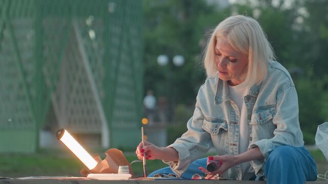 close up portrait of painter seated on bench in twilight park painting detailed strokes under lamp shine with focused expression while children play nearby framed by trees and jars of paint