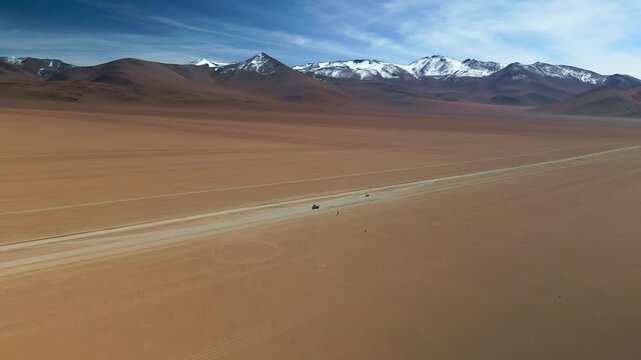 Drone flying forward over Dali Desert with camera tilting down, jeep parked on dirt road with people nearby, snow-capped mountains and blue sky background.