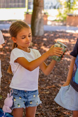 Young girl with long hair wearing white shirt and denim shorts, joyfully shaking a glass jar filled with nature treasures, surrounded by classmates in an outdoor learning environment