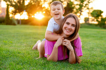 Fototapeta premium mother and child are walking and playing on a green lawn in the summer, having fun together and lying on the grass. The mother and son are hugging and kissing, laughing and smiling.