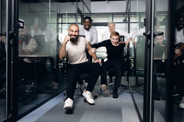 Multiracial colleagues engage in playful office chair race down modern corridor with glass walls. energy and teamwork are highlighted as they enjoy lively break from work tasks.