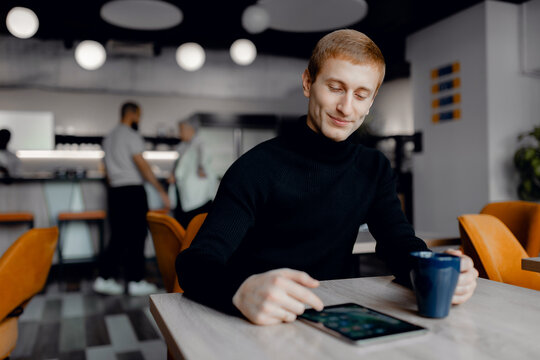 Young redhead man in modern cafe setting, interactively using tablet while sipping coffee, surrounded by warm lighting and contemporary interior design, capturing moment of leisure.