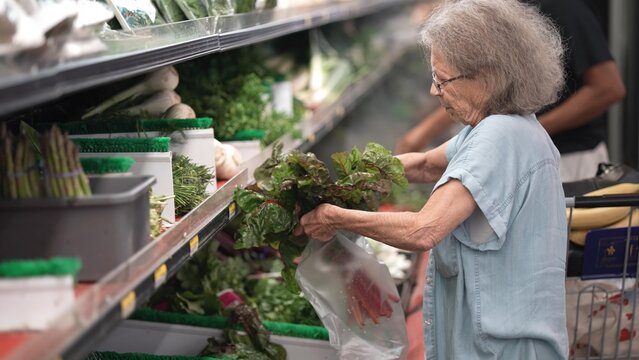 An elderly woman carefully selects fresh vegetables in a grocery store. She is focused on choosing healthy food options while enjoying her shopping experience.