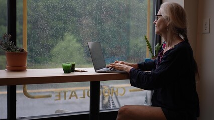 Portrait of a smiling mature woman happily drinks coffee while working on her laptop and phone in a warm cafe on a rainy day, surrounded by a peaceful atmosphere.