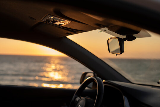 A car interior with a view of the ocean at sunset. The steering wheel and rearview mirror are visible. The scene conveys a sense of tranquility and freedom.