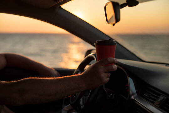 A man holds a coffee cup while driving a car by the sea at sunset. The scene conveys relaxation and freedom with ample copy space.