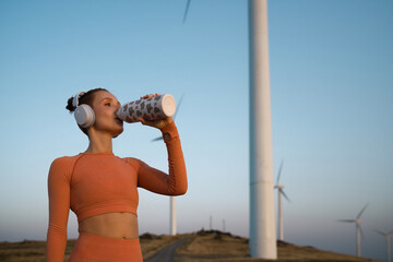 Young sportswoman drinking water from bottle near wind turbines during sunset