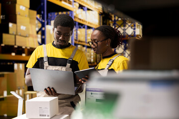 Warehouse workers overseeing parcels tracking info on laptop, preparing pallets for outbound shipment. Organized depot with storage racks, cardboard boxes and inventory processing tools.