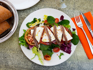 Fresh salad with tomato, cheese and anchovies on white plate, served with bread.
