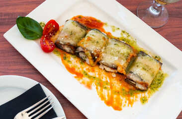 Eggplant rolls stuffed with cheese filling are laid out on plate. Dish is decorated with spinach leaves, cherry tomatoes and richly flavored with pesto sauce