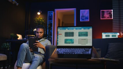 Black remote worker navigating on tablet next to screen running analytics, sitting at home working on his daily tasks with multiple devices. Leisure and job productivity in loft. Camera A.