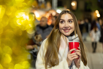 In middle of noisy crowd at holiday fair, there is dreamy girl with glass of warm tea in her hands. Strolling tourist dreamily examines Christmas goods and enjoys atmosphere