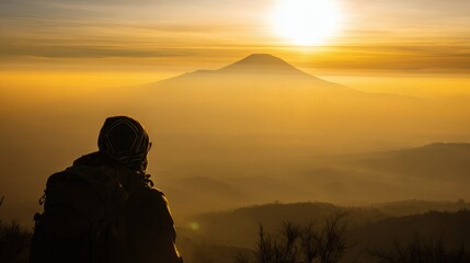 A mountain climber gazes at a distant peak, silhouetted against a golden sunrise in a dramatic landscape.